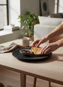 Paniery handwoven black raffia and sisal flat plate with concentric circle design, holding bread slices and nuts.