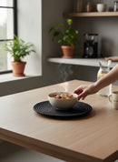 Paniery black woven round placemat with concentric circle design, holding a bowl of cereal.