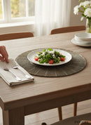 Paniery round beaded placemat in bronze with a sunburst pattern, set on a wooden table with a salad plate.