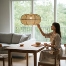 Paniery handwoven natural raffia lampshade with an organic shape, casting a warm diffused light. A woman in a neutral dress reaches towards it in a modern dining room.