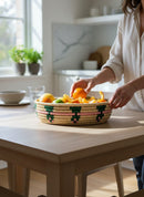 Paniery oval handwoven basket with pink and green geometric pattern, filled with fruit on a wooden table.