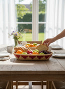 Paniery oval handwoven basket with red geometric pattern, filled with fruit and bread on a wooden table.