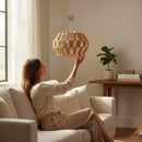 Paniery handwoven natural raffia pendant lampshade with a spherical, open-weave design, held by a woman sitting on a sofa.