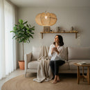 Paniery handwoven natural raffia lampshade with an organic shape, creating diffused light. A woman sits on a sofa nearby.