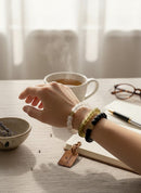 Paniery African Heritage recycled glass bracelets in white, olive green, and black, featuring a spiral charm. Stacked on a wrist with a cup of tea and notebook.
