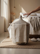 Paniery Senegal handwoven laundry basket in natural tones with leather handles, featuring a lid and a draped cloth.