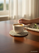 Paniery handwoven white coaster with a textured weave, protecting a coffee cup on a wooden table.