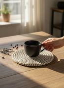 Paniery handwoven white round coaster made of natural fibers, protecting a wooden table from a black coffee mug.
